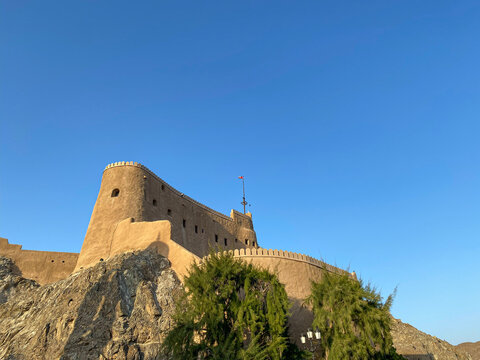 Majestic ancient fort perched high on rugged rocks under a clear blue sky in Mutrah Fort, Muscat, Oman. The stone walls and imposing circular towers showcase historical architecture and strength.