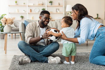 Happy black family enjoys quality time together in their living room. The father and mother engage their curious son, playing with a ball on a cozy carpet, creating joyful memories.