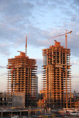 Two tall buildings under construction with cranes in the background. The sky is cloudy and the sun...