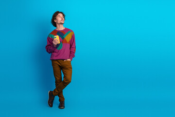 Young stylish man with curly hair holds yellow phone against blue background in colorful sweater and brown pants