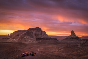 View of the sun ablaze, painting the sky with fiery hues over the rugged, arid landscape and distant mesas, a lonely vehicle sitting on the plain, Aktau, Mangystau Region, Kazakhstan.
