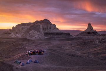 View of tents clustered on the arid ground under a vibrant sunset sky, shadowed by towering rock formations, Aktau, Mangystau Region, Kazakhstan.