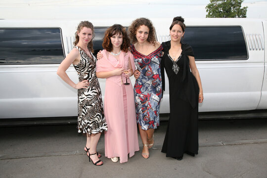 Four women in fancy dresses pose for a picture in front of a white limousine. Scene is elegant and sophisticated, as the women are dressed up for a special occasion