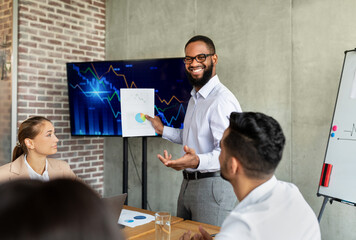 A black male entrepreneur presents company growth data during a corporate meeting in an office. He engages coworkers with a financial report while standing next to a monitor, smiling confidently.