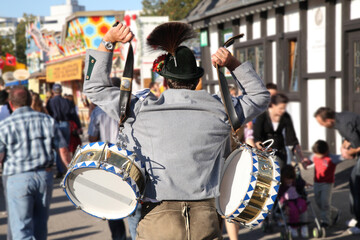 A man wearing a hat and holding two drums is surrounded by a crowd of people. The atmosphere is...
