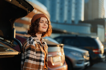 Woman by car in parking city, smile near open trunk in warm sunlight at golden hour glow....