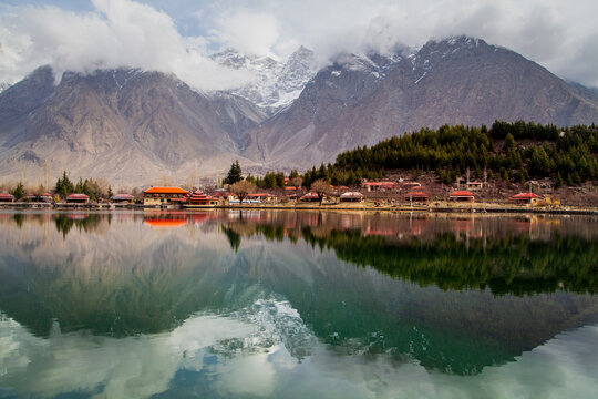 View of the serene lake reflecting jagged, snow-dusted mountains and a quaint village nestled against the forested hillside in Kachora, Gilgit Baltistan, Pakistan.