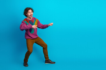 Young man with curly hair in a colorful sweater poses playfully against a blue backdrop for lifestyle fashion and advertising imagery