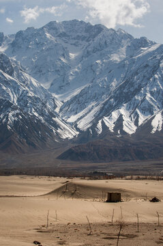 View of a lone shack standing against a vast, beige desert contrasting with the massive, snow-capped mountains under a pale sky, Skardu, Gilgit Baltistan, Pakistan.