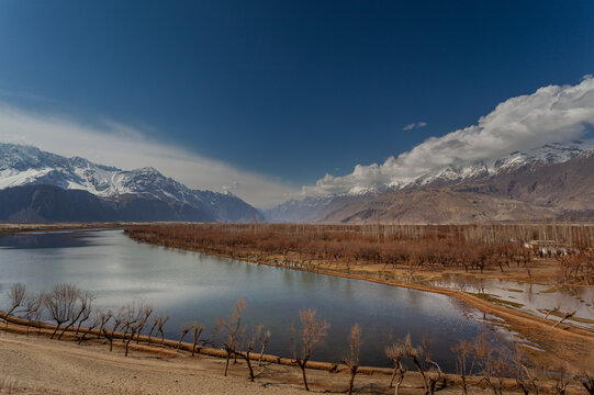 View of serene waters reflecting the clear blue sky, bordered by barren trees and snow-capped mountains under the crisp sunlight, Skardu, Gilgit Baltistan, Pakistan.