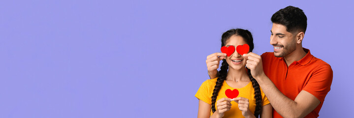 A young couple stands close together, playfully holding red heart-shaped cutouts in front of their eyes. They smile with happiness, showcasing their affection against a light purple backdrop.