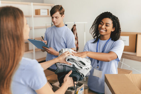 A group of volunteers sorts through donated clothes at a community center. One woman smiles while handing items to another as a young man takes notes for inventory. - Powered by Adobe