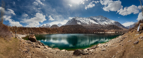 View of shimmering turquoise lake reflecting the snow-capped mountains under a vast sky with scattered clouds, Kachura, Skardu, Pakistan.