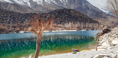 View of a serene lake reflecting the rugged mountains and bare trees under a pale sky, with a small boat resting on the shore, Kachura, Skardu, Pakistan.