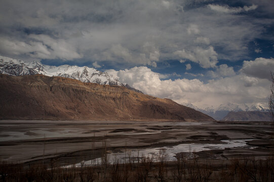 View of rugged, brown mountains meeting a flat, barren riverbed under a sky streaked with dramatic clouds, snow-capped peaks in the distance, Skardu, Gilgit Baltistan, Pakistan.