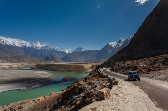 View of a blue jeep winds along a stark road, paralleling a vibrant river against a backdrop of towering, snow-capped mountains, Skardu, Gilgit Baltistan, Pakistan.