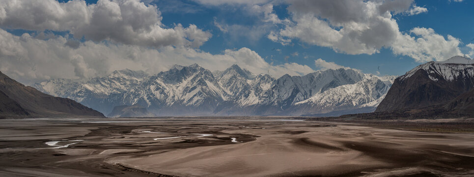 View of stark desert landscape with winding streams contrasts sharply against the majestic snow-capped mountains under a dynamic sky, Skardu, Gilgit Baltistan, Pakistan.