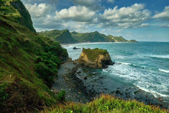 View of the rugged coastline with lush green cliffs meeting the turquoise sea under a sky brushed with clouds in Pantai Menganti, Kebumen, Central Java, Indonesia.