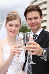 A bride and groom are holding up their champagne glasses to toast their wedding. The bride is wearing a white dress and the groom is wearing a suit. Scene is celebratory and happy