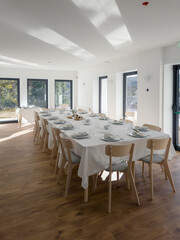 A bright elegant dining hall is prepared for a festive group meal, with a long white table set with plates, glasses and napkins, surrounded by chairs and large windows that fill the room with light