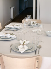 Closeup of an elegant festive dining table shows neatly arranged plates, glasses and white napkins tied with rustic twine and rosemary sprigs, highlighting refined details in a bright modern meal room