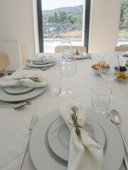 Closeup of an elegant festive dining table shows neatly arranged plates, glasses and white napkins tied with rustic twine and rosemary sprigs, highlighting refined details in a bright modern meal room