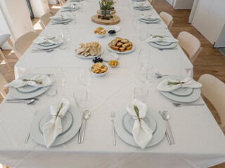 A long table in a bright dining room is set for a group gathering, with plates, glasses and a variety of savory appetizers such as bread, meat croquettes, sausage toast, olives and lupin beans arrange