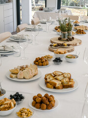 A long table in a bright dining room is set for a group gathering, with plates, glasses and a variety of savory appetizers such as bread, meat croquettes, sausage toast, olives and lupin beans arrange