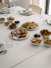 A long table in a bright dining room is set for a group gathering, with plates, glasses and a variety of savory appetizers such as bread, meat croquettes, sausage toast, olives and lupin beans arrange