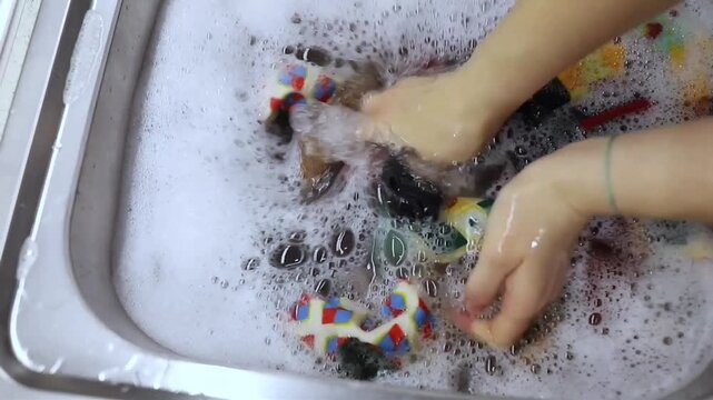 Female hands scrubbing colorful patterned socks under foam water inside metal sink, bright fabric tones visible through soapy surface, manual laundry action showing grip, splash, texture contrast, wet