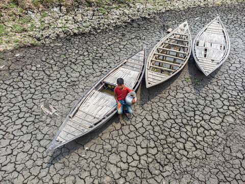 Rajshahi, Bangladesh - 09 May 2024: Aerial view of cracked earth cradling stranded wooden boats, a stark tableau of drought's grip, with a lone figure bearing a water vessel.