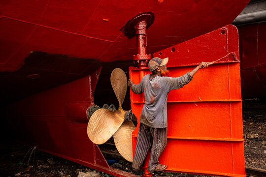Dhaka, Bangladesh - 06 May 2024: View of a worker diligently painting the bright red hull of a ship, contrasting against the dull metallic propeller and the worker's worn attire.