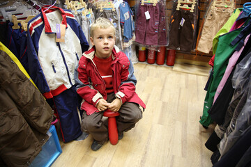 A boy is sitting on a stool in a store with clothes hanging on racks. The boy is looking at the...