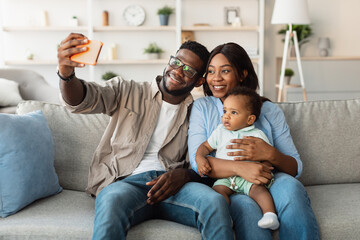 A joyful African American family enjoys quality time at home. The dad, mom, and their happy baby smile as they take a selfie on the couch in their comfortable living room.