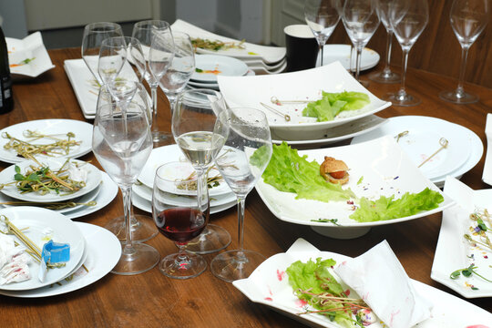 Leftover food and dirty dishes scattered across a wooden table, featuring empty wine glasses and plates, creating a post-event atmosphere of culinary indulgence and celebration