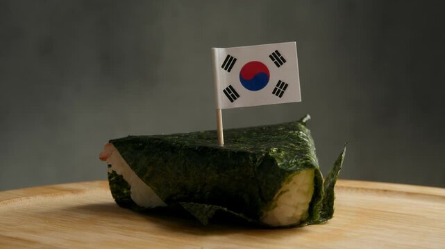 Close up shot of rice triangle wrapped in seaweed with South Korean flag on toothpick, scene highlights Korean gimbap style snack made from local rice, nori and fillings used in everyday Korean
