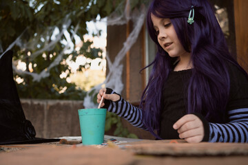 Little girl wearing purple wig and witch costume smiling happily and painting by paint and brush