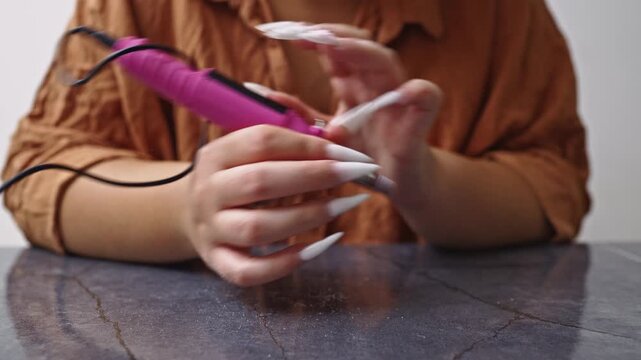 A close-up shot of a woman carefully using a pink electric nail file to shape and refine her long, white artificial nails, emphasizing beauty and personal care.