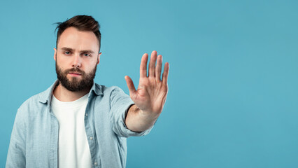 A man with a beard and short hair stands against a solid blue background. He raises one hand,...