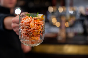 A person holds a clear glass filled with cooked pink shrimp or prawns, garnished with a lemon wedge and fresh dill, in a restaurant or bar setting with a blurred background
