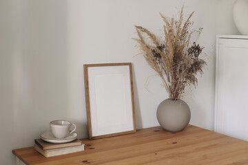 Minimal cozy interior scene with blank wooden picture frame mockup. Dried flowers, grass in vase. Coffee cup, books on table. Neutral Scandinavian home decor, natural rustic styling. Lifestyle design