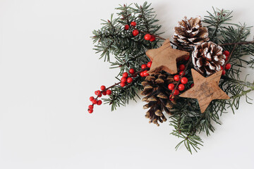 Christmas festive still life. Decorative composition. Pine cones, red holly berries, wooden stars, fir tree branches isolated on white table background. Winter floral flat lay, top view. Copy space