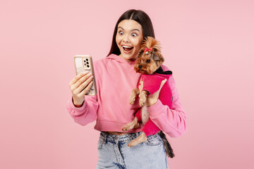 Young woman holding her pet dog and smartphone, surprised by the result of a selfie