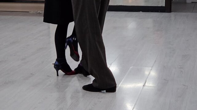 Man and woman are dancing tango on a wooden floor. The man is wearing a suit and the woman is wearing a black dress