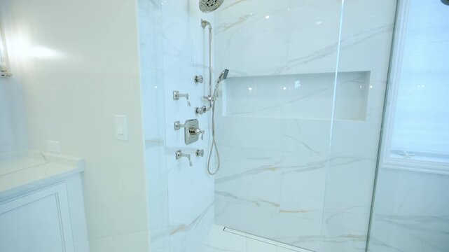 Clear glass shower door in a bright, modern bathroom. The background reveals white cabinetry, polished marble tile, and high-end shower fixtures