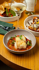 A beautifully plated fish dish lies in a shallow bowl, surrounded by colorful vegetables and sauce. A crisp glass of white wine adds elegance, while the warm restaurant ambiance invites enjoyment