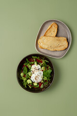A colorful bowl of salad features fresh greens, sliced cucumbers, and creamy poached eggs, complemented by slices of golden toasted bread on a soft green surface