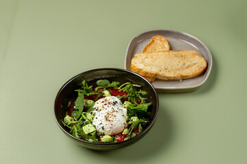 A vibrant bowl of salad features a perfectly poached egg atop fresh greens, with sliced tomatoes and small vegetables. There's a side of toasted bread, all displayed on a soft green table