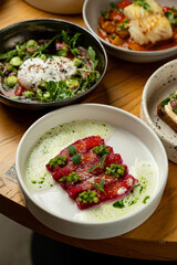 A vibrant dish presents thinly sliced meat topped with herbs and sauces, surrounded by additional creative plates filled with fresh greens and savory delights