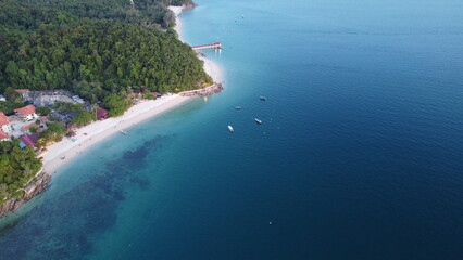 Drone image of tropical beach with pier, anchored boats, red-roof buildings, forest, and clear water. © Na'im